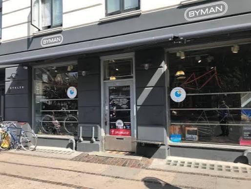 Mørkegrå butiksfacade med et hvidt BYMAN-logo i en rund ramme og teksten CYKLER i mindre bogstaver under – indgangen til Byman Cykler på Øster Farimagsgade.
